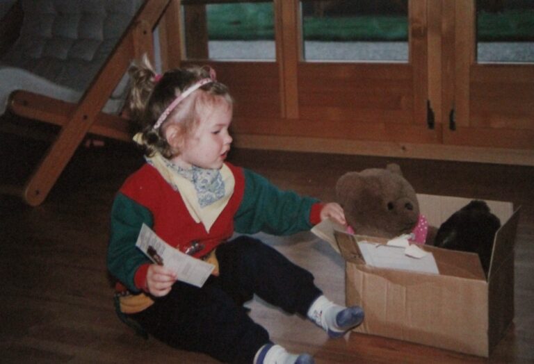 Michelle als Kind, wie sie ihrem Teddybär in der Kartonschachtel ein Büchlein vorliest.