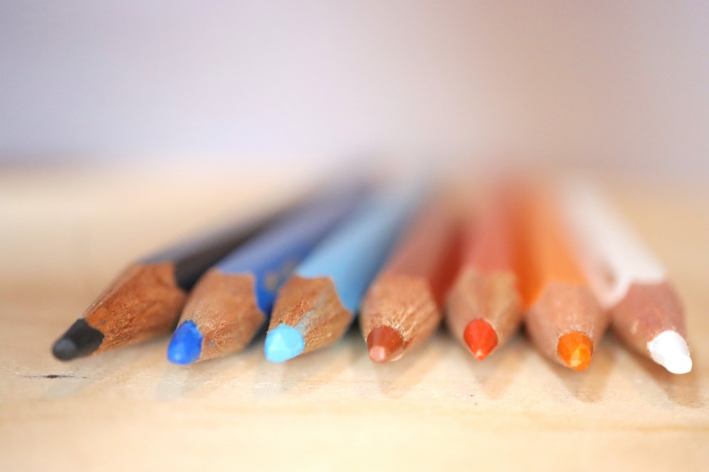 Buntstifte in Schwarz, Blau, Braun, Orange und weiss liegen auf einem Holztisch. Das sind die Unternehmensfarben von Buchfink-Design